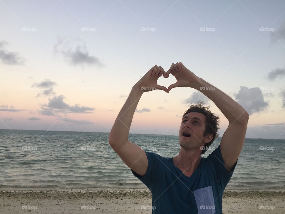 Man at the beach in Mauritius forming a heart broth his hands above his head. It is evening and the sun is low.  