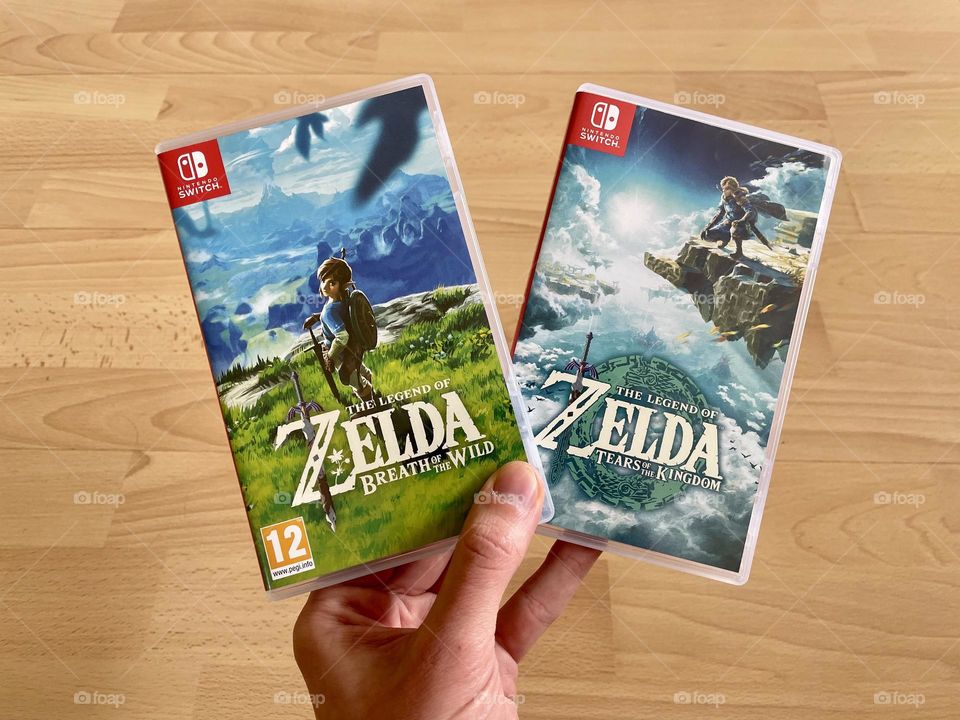 The Legend of Zelda: Tears of the Kingdom and Breath of the Wild cases held by a man's hand on wooden background.