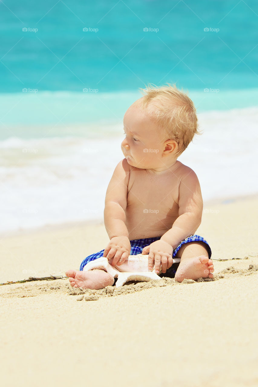 A Brunette Haired Toddler Playing In The Soft Sand And Looking At The Clear Blue Waves,have a nice day.