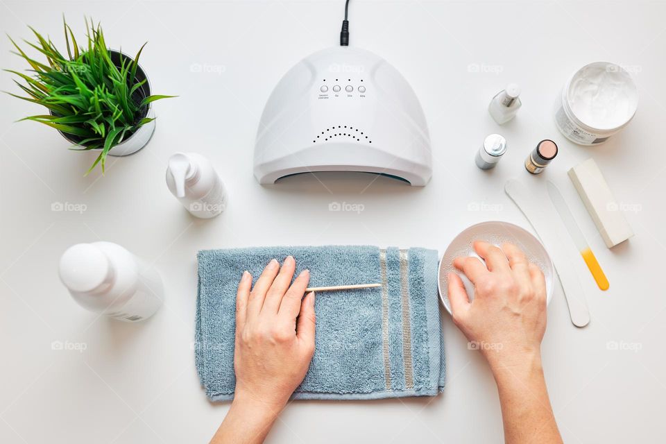 Woman preparing nails to apply gel hybrid polish using UV lamp. Beauty wellness spa treatment concept. Cosmetic products, UV lamp, green leaves on white table. Spa, manicure, skin care concept. Flat lay, overhead view