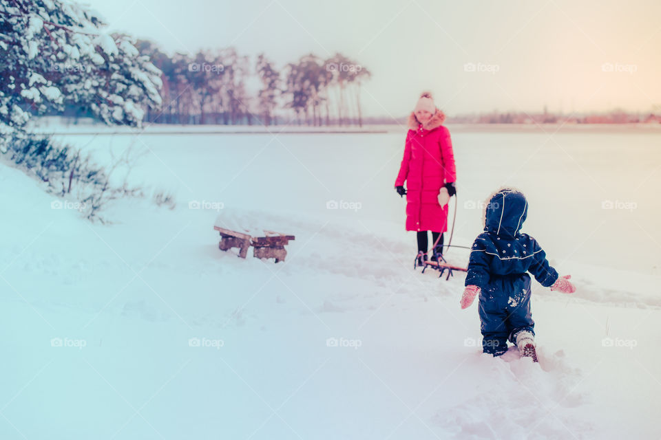 Mother and her little daughter are spending time together walking outdoors in forest in winter while snow falling. Woman is pulling sled, a few years old girl is walking through the deep snow, enjoying wintertime