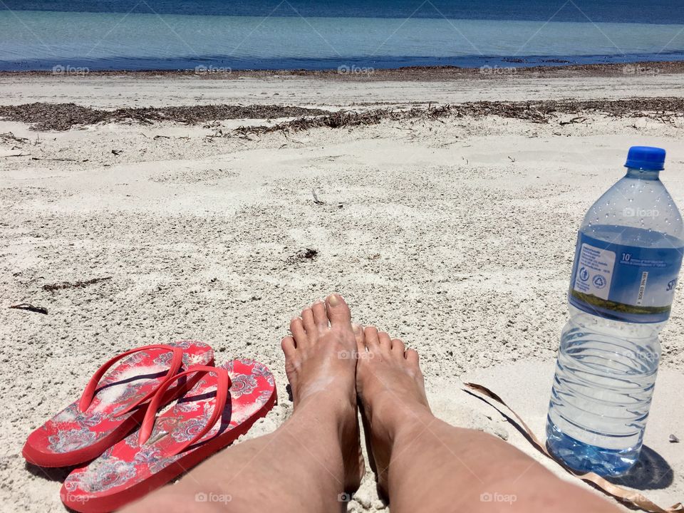 Tanned legs sandy feet woman laying on secluded beach with water bottle and sandals flip flops thongs by her side taking in the view 