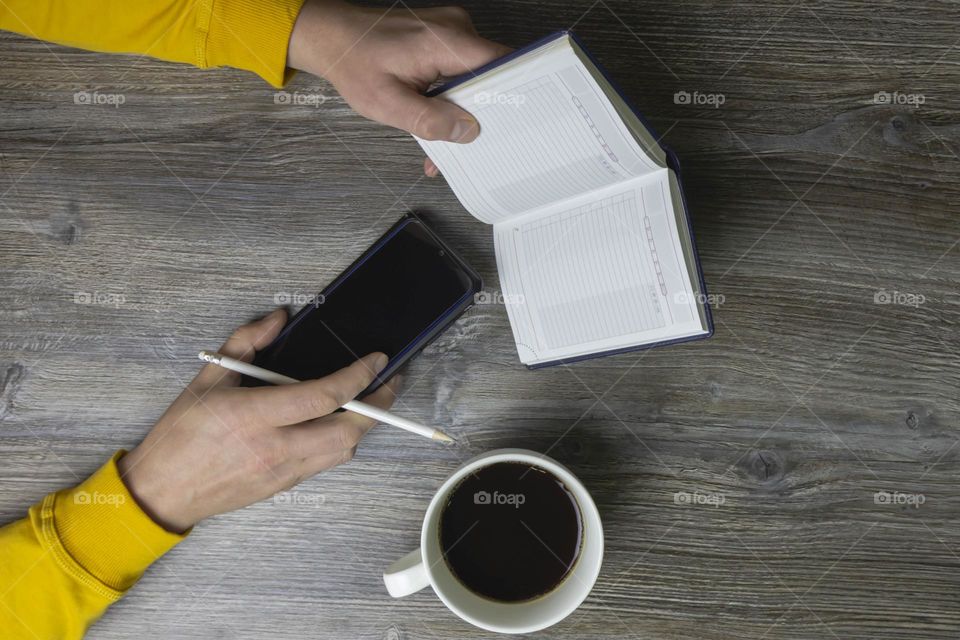 A man drinks coffee in the morning, works on the phone and makes notes in pencil in a notebook. Men's hands hold a white cup of coffee and a mobile phone in a yellow jacket. The hands lie on a gray wood background.