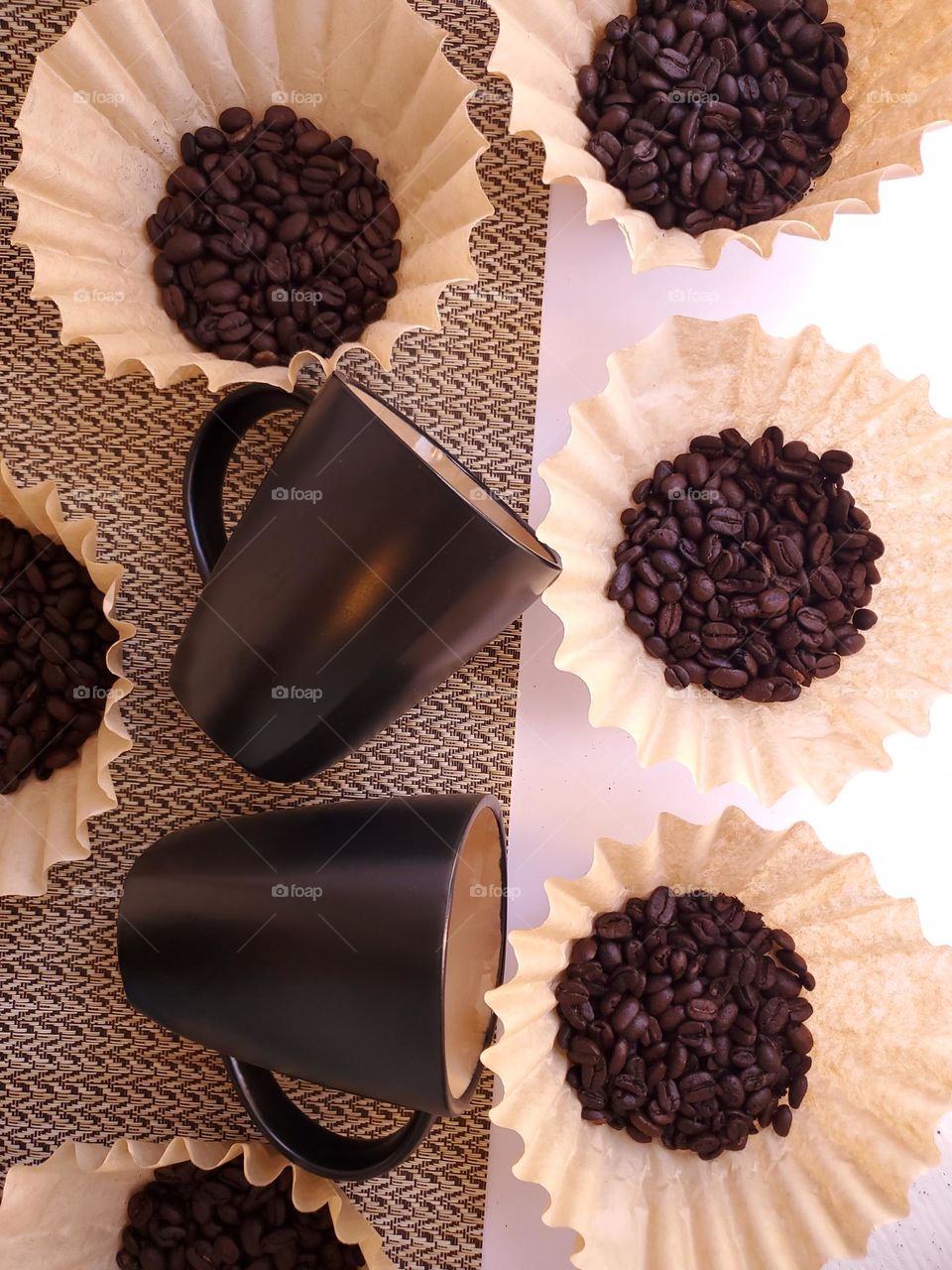 Coffee beans displayed in coffee filters surrounding two black coffee cups on partial white and partial beige surface.