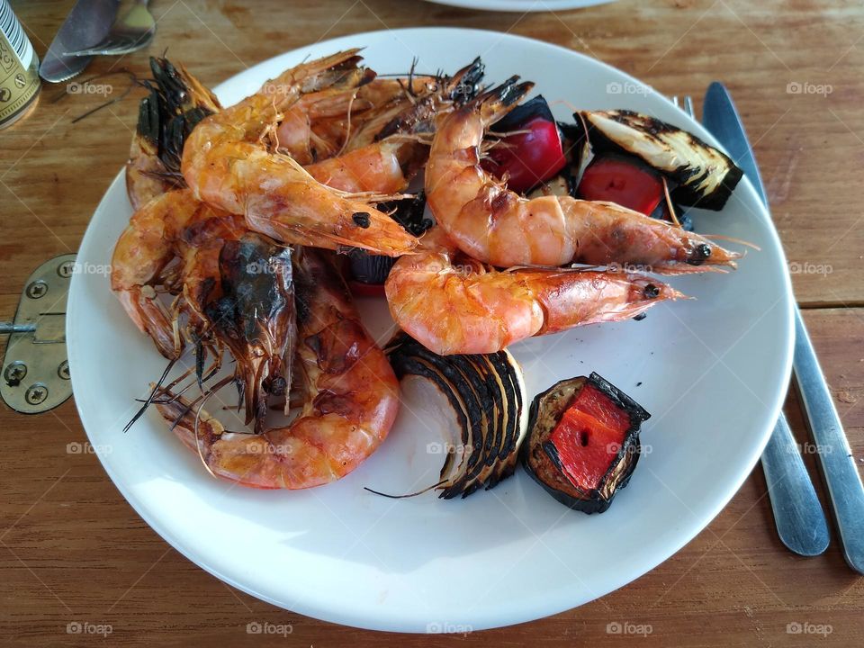 A delicious looking meal of Large Grill Cooked Pink Coloured Whole Prawns in their Shells, with Red Bell Peppers and White Onions on a White Plate on a Wooden Table, with a Knife and Fork and the Silver Hinges of the Folding Table showing.