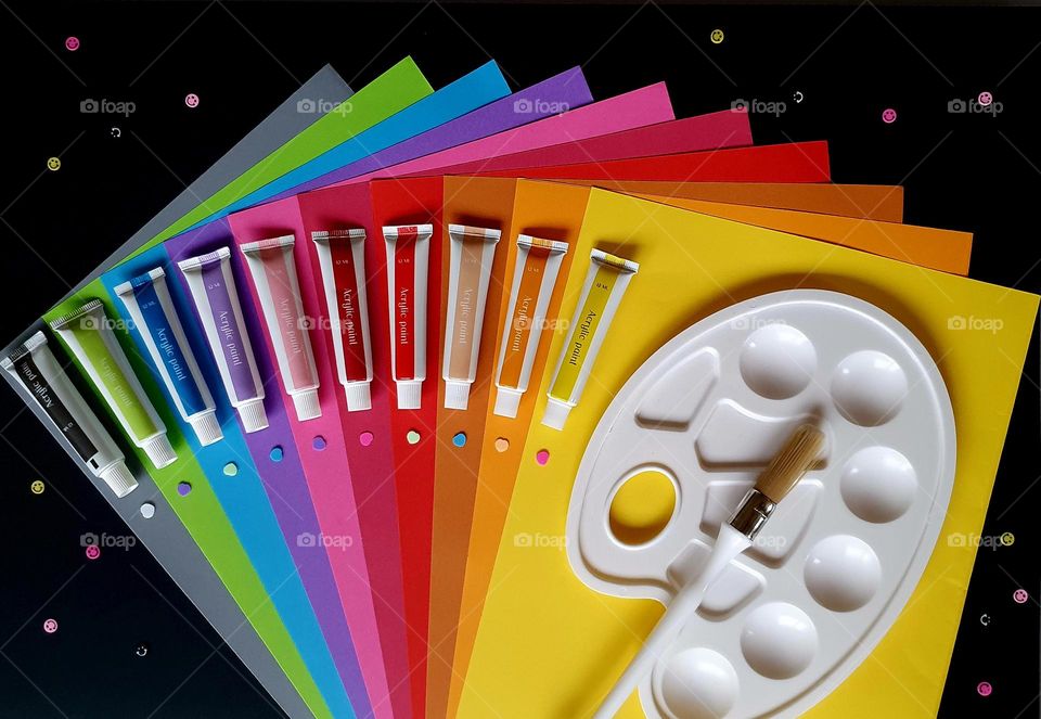 Colourful flatlay of small acrylic paint tubes placed on a fan-shaped set of colour papers with a black background, together with a paint brush and paint pallet
