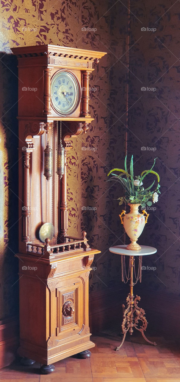 Old fashioned grandfather clock with pendulum and antique vase with flowers in a room with retro designer wallpaper.