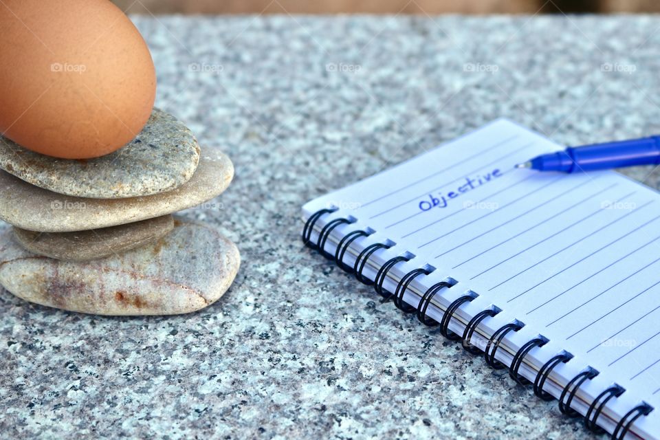 Whole intact hen’s chicken brown egg balancing atop smooth stone rock stack on marble surface, with spiral notebook and objective handwritten title, concept business, motivational, or life applications, with copy space