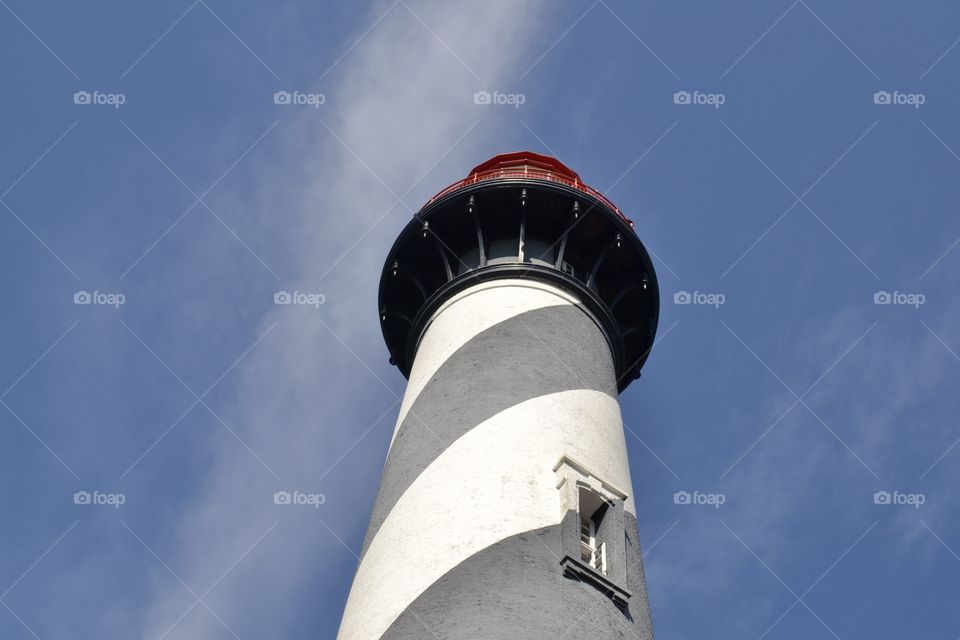 Looking upward to the top of a black and white striped lighthouse with a circular red top against a bright blue sky with white puffy clouds