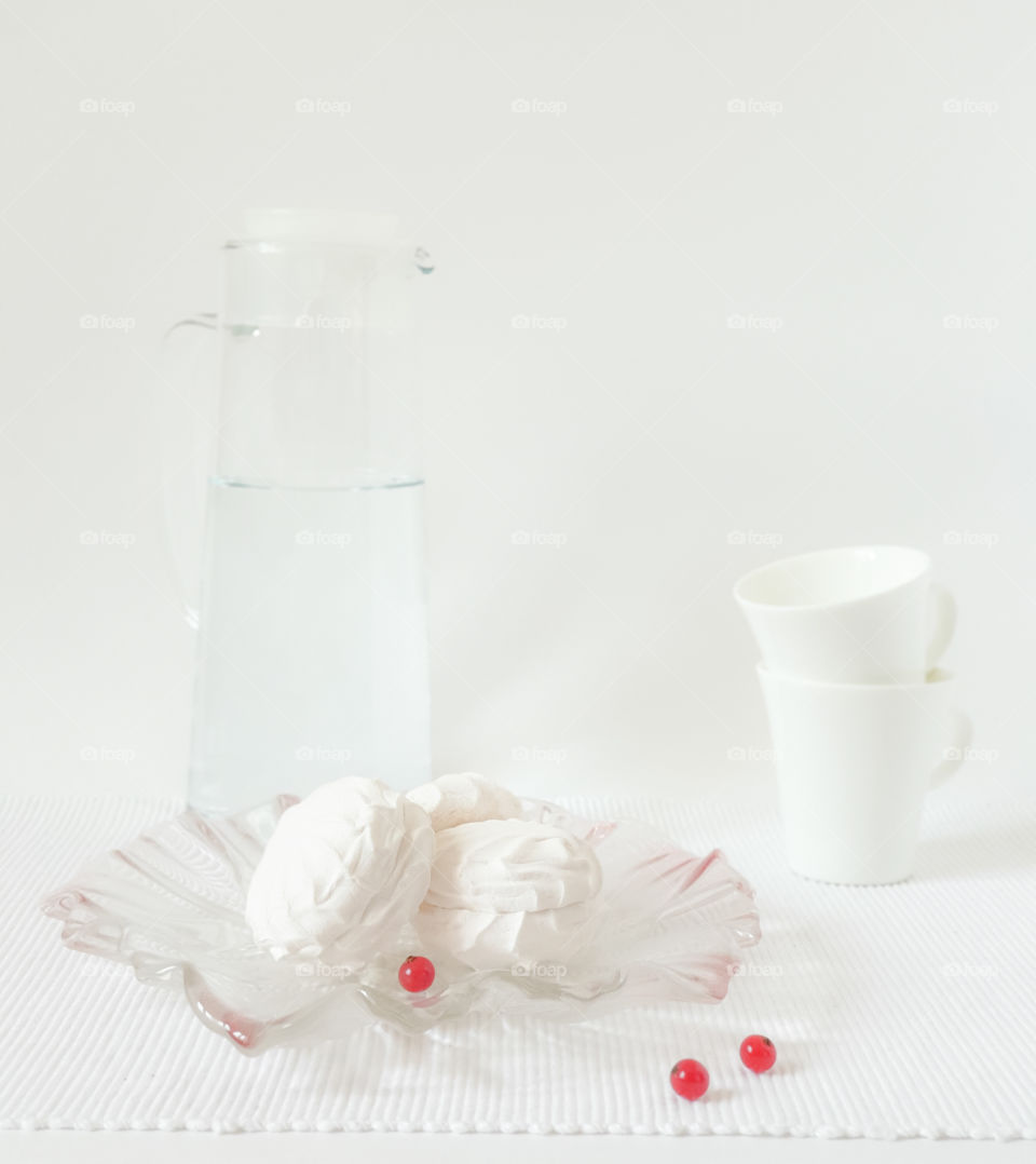 Still life in a rustic style with pastries.  White marshmallow with red currant berries, tea cups and a glass jug of water on a wooden board and textured white napkin.  Tasty snack