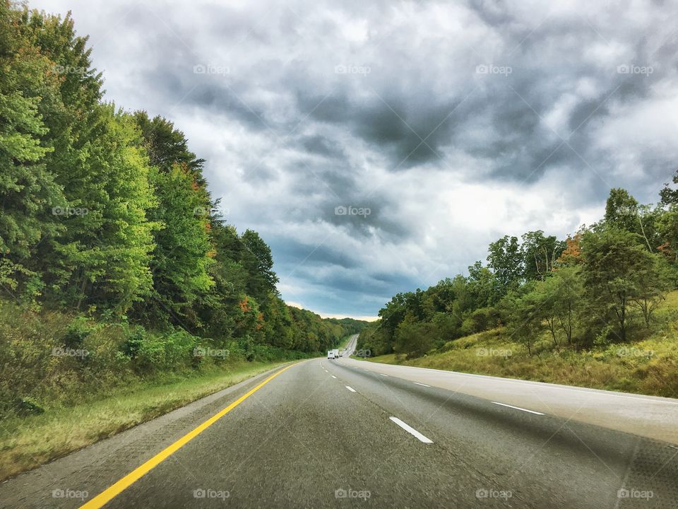 A divide interstate highway lined with vibrant green trees under a sky filled by ominously gray cloud. 