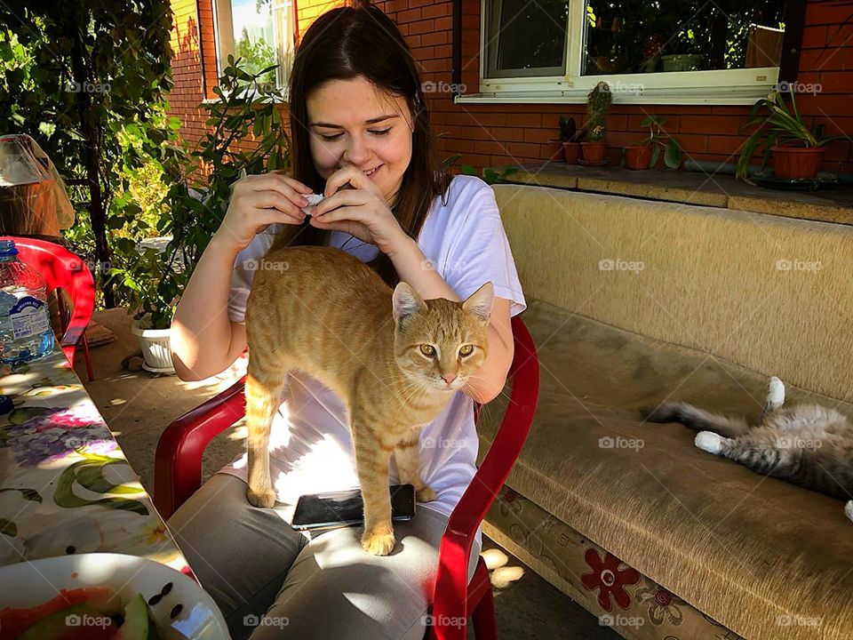 Camping.  A teenage girl sits on a chair and smiles at a ginger cat that stands on her feet.