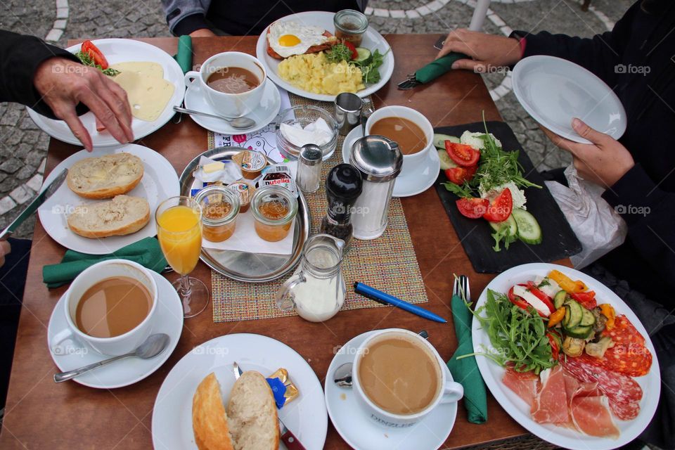 Top view of a hearty breakfast with sausage, cheese, rolls, sweets, coffee and vegetables together with friends 