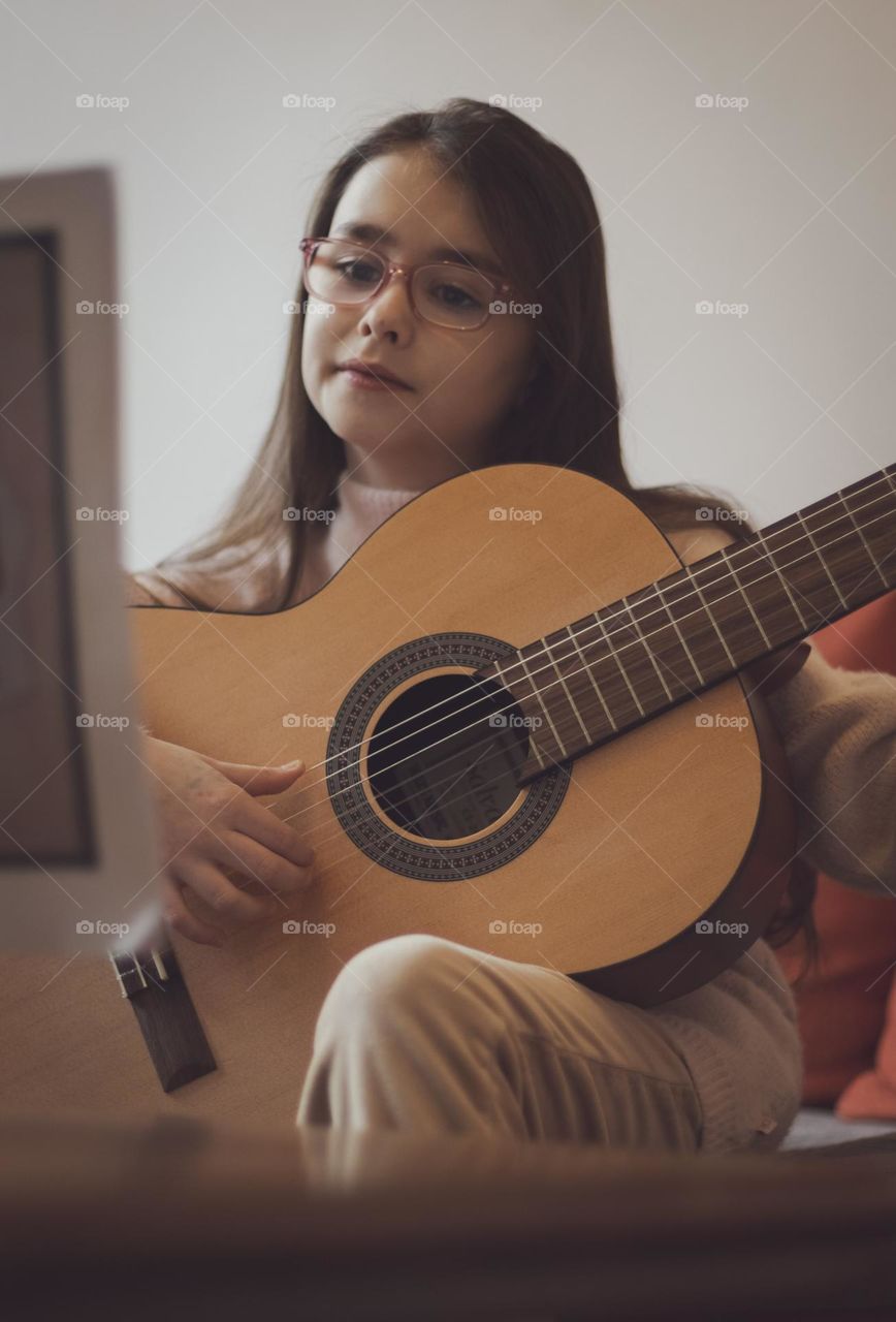 Portrait of one beautiful caucasian girl in glasses with long flowing hair with a guitar in her hands sits on a sofa at a table with notes and learns them, doing music at home, close-up side view. Music education concept.