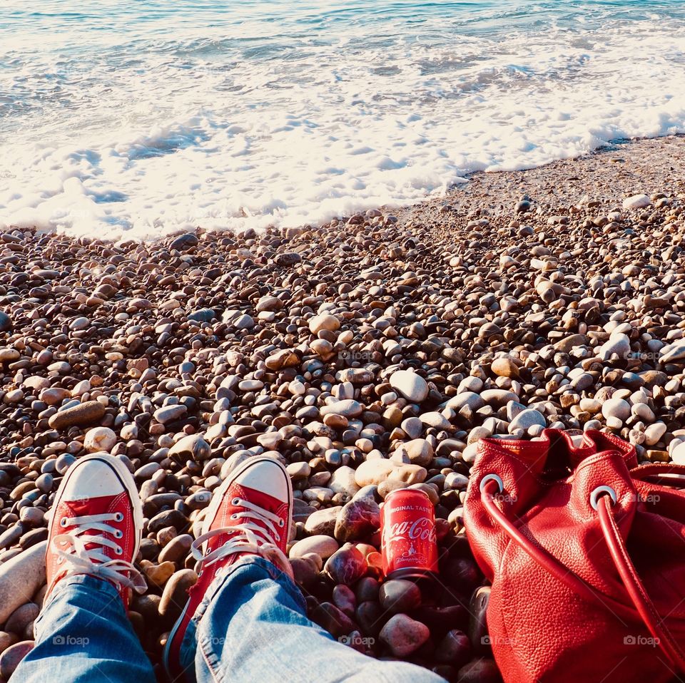 A can of Coca Cola on the beach with red sneakers and red leather bag.