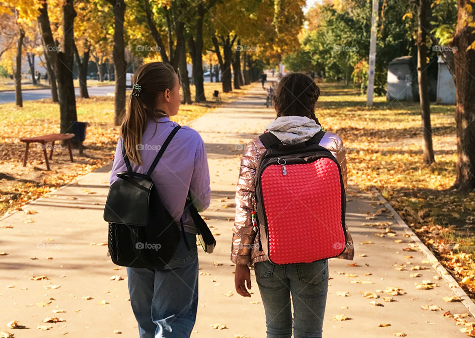 Two girls students with backpacks walking by the autumn city road 