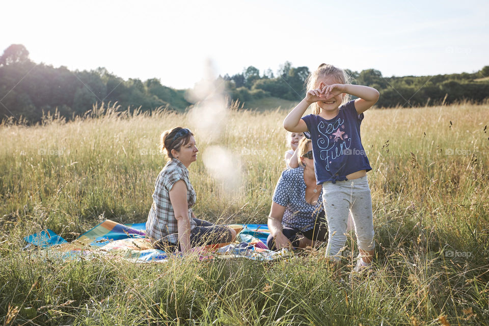 Little girl making a hand gesture for taking picture. Family spending time together on a meadow, close to nature. Parents and kids sitting on a blanket on grass. Candid people, real moments, authentic situations