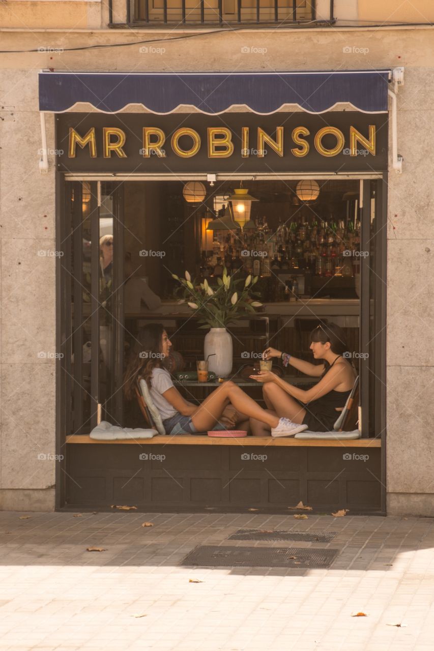 Two girls sitting in a window seat at a local bar in barcelona, talking with each other and having a relaxed great time