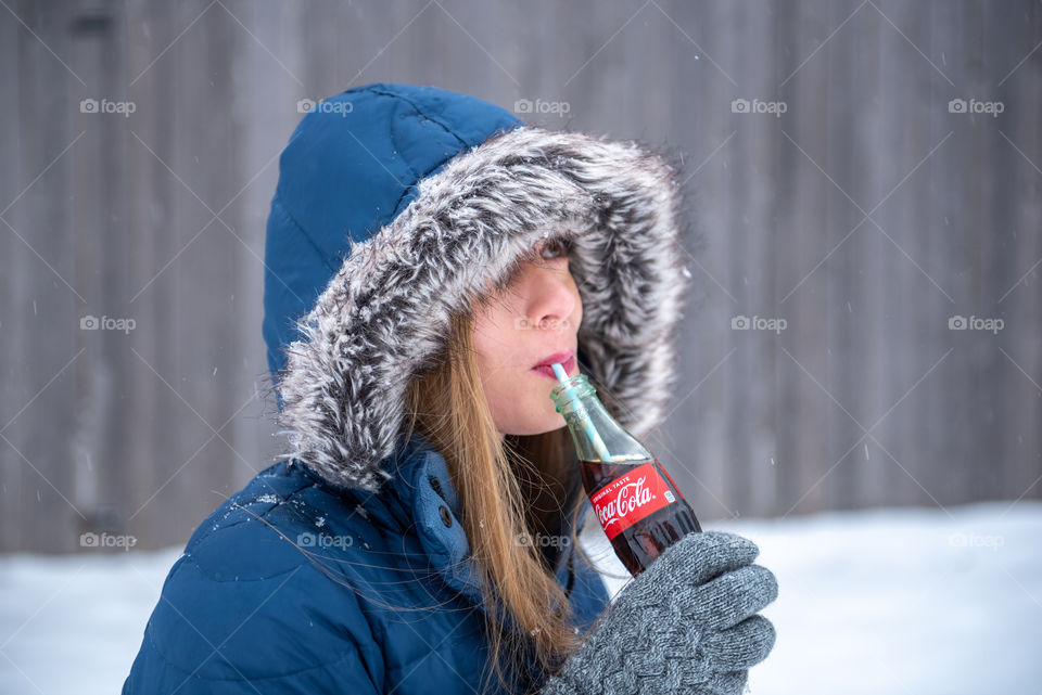 Millennial woman in a winter coat drinking a bottle of Coca-cola from a straw outdoors in the snow