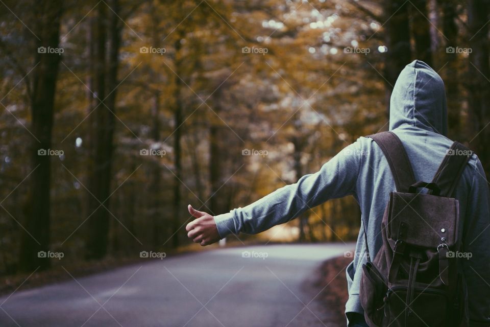 A hooded hitchhiker with backpack extending in thump on the side of an empty road