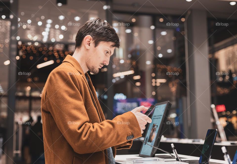 Portrait of a young handsome Caucasian brunette guy in a brown coat in a hardware store with a smartphone in his hands examining him, close-up side view.