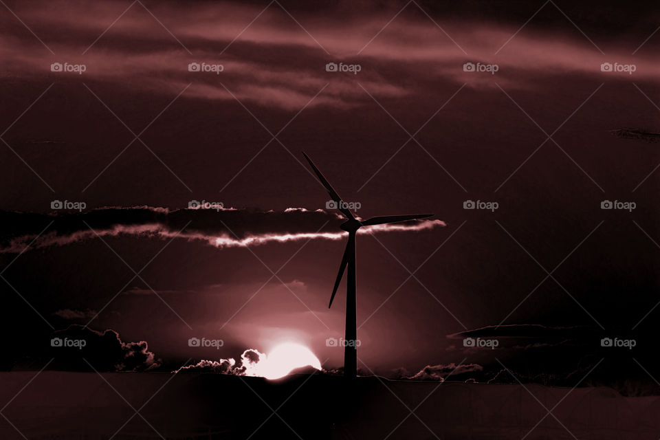 Wind turbine on the horizon at sunset. Dark red sky with bright sun shape behind a cloud. Picture taken in Salento, Puglia, Italy.