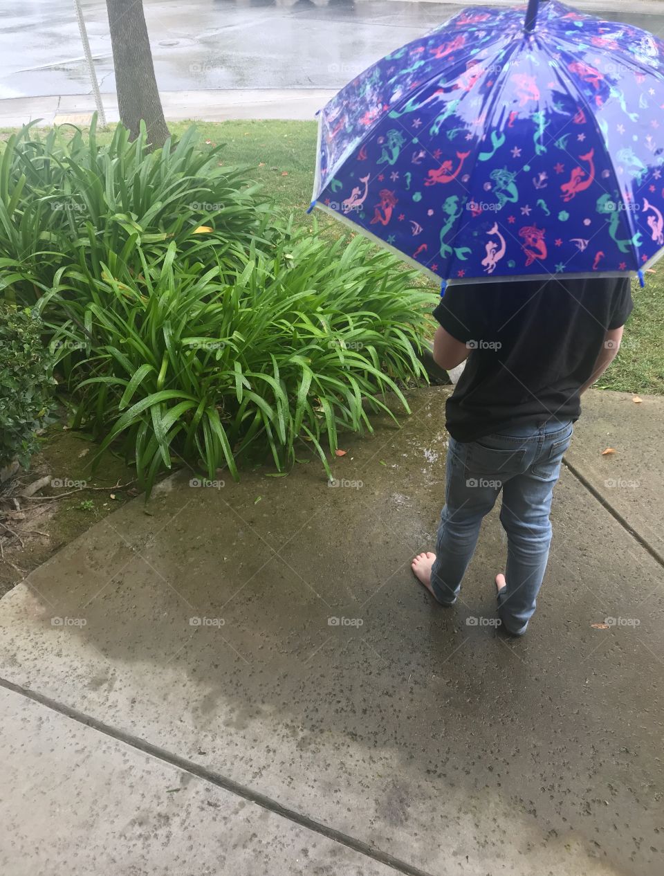Little boy standing in the rain with a blue umbrella next to the green grass outside. little feet in the ☔️