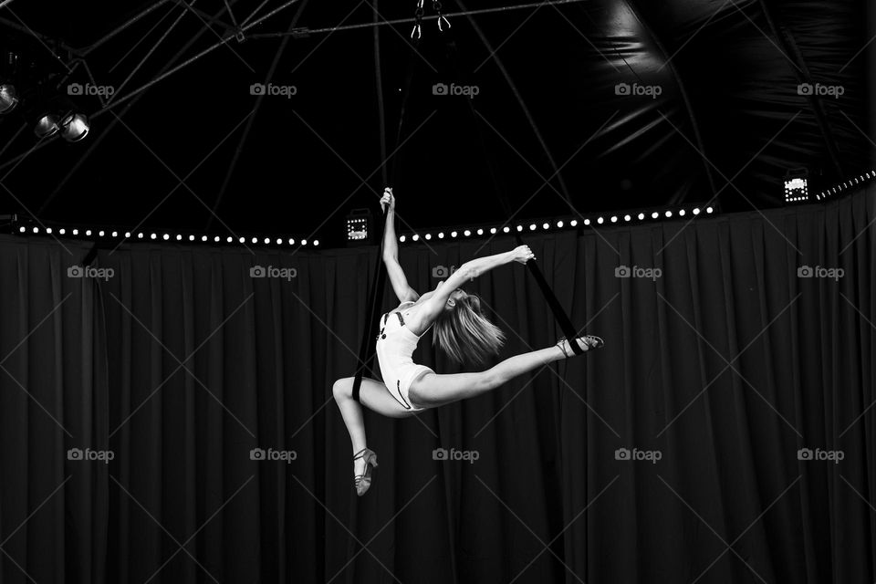 A black and white portrait of a female circus artist doing acrobatic stunts on wires in a white outfit in a circus tent.