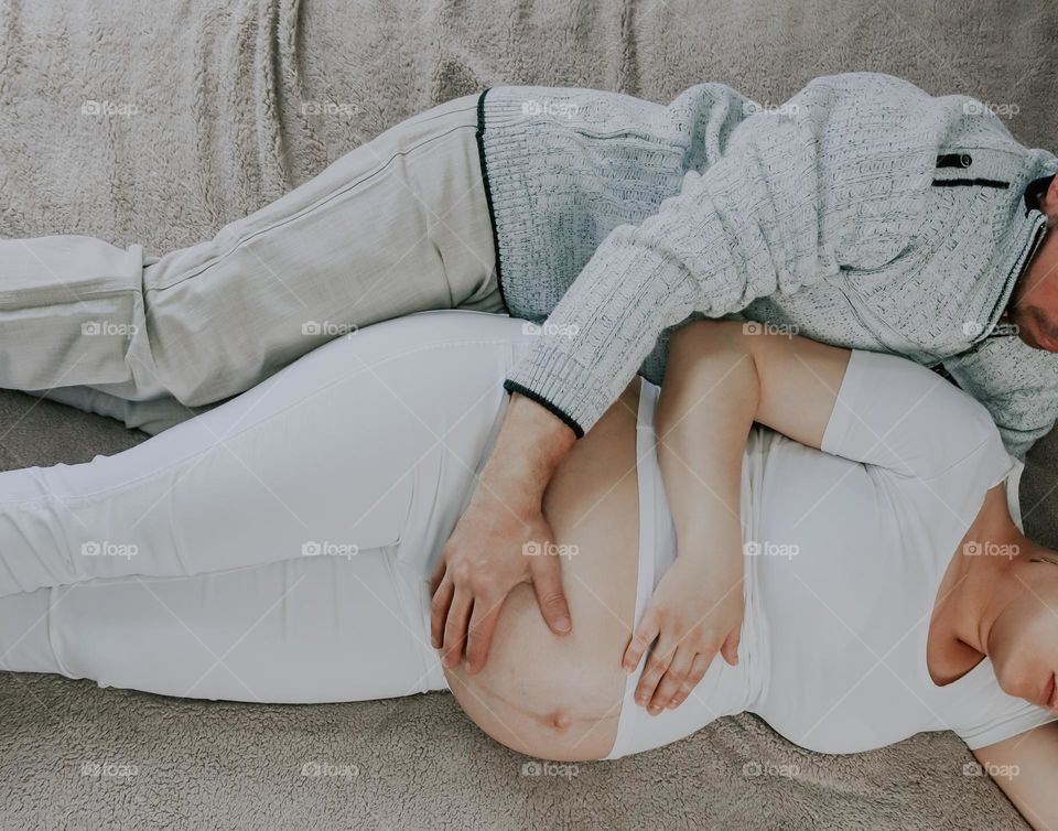 A young caucasian guy hugs the belly of his pregnant wife while lying on the bed in the room, flat lay close-up. The concept of a married couple, romantic pastime, expectation of a child, mental support.