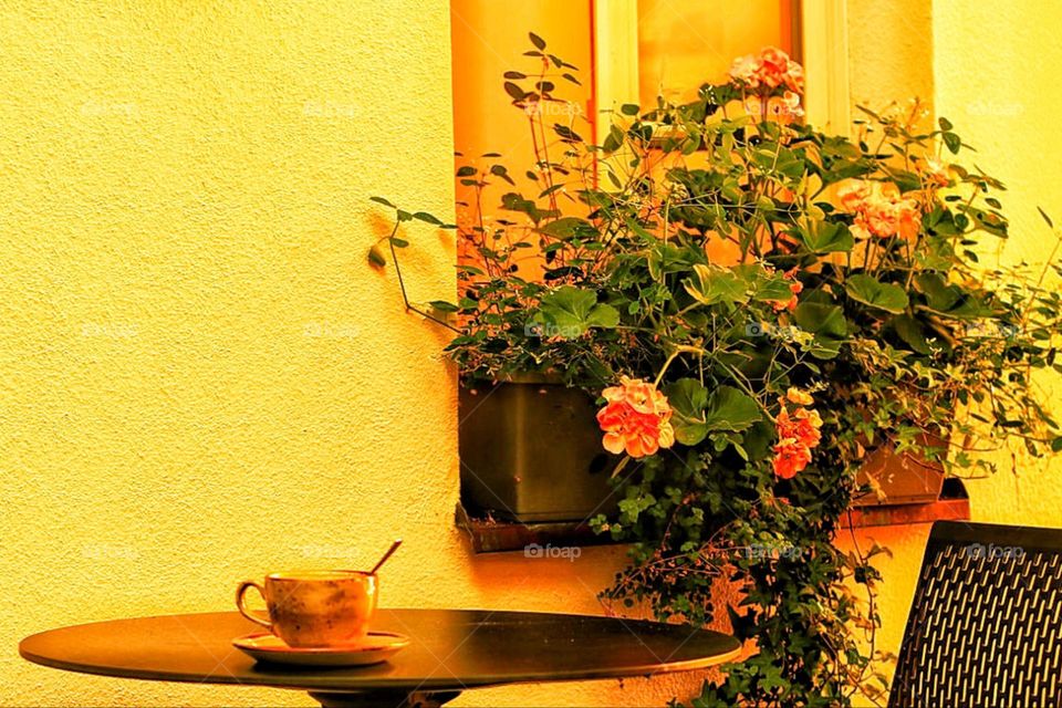 A cup of coffee is on a round table next to flowers in the city in front of a yellow house 