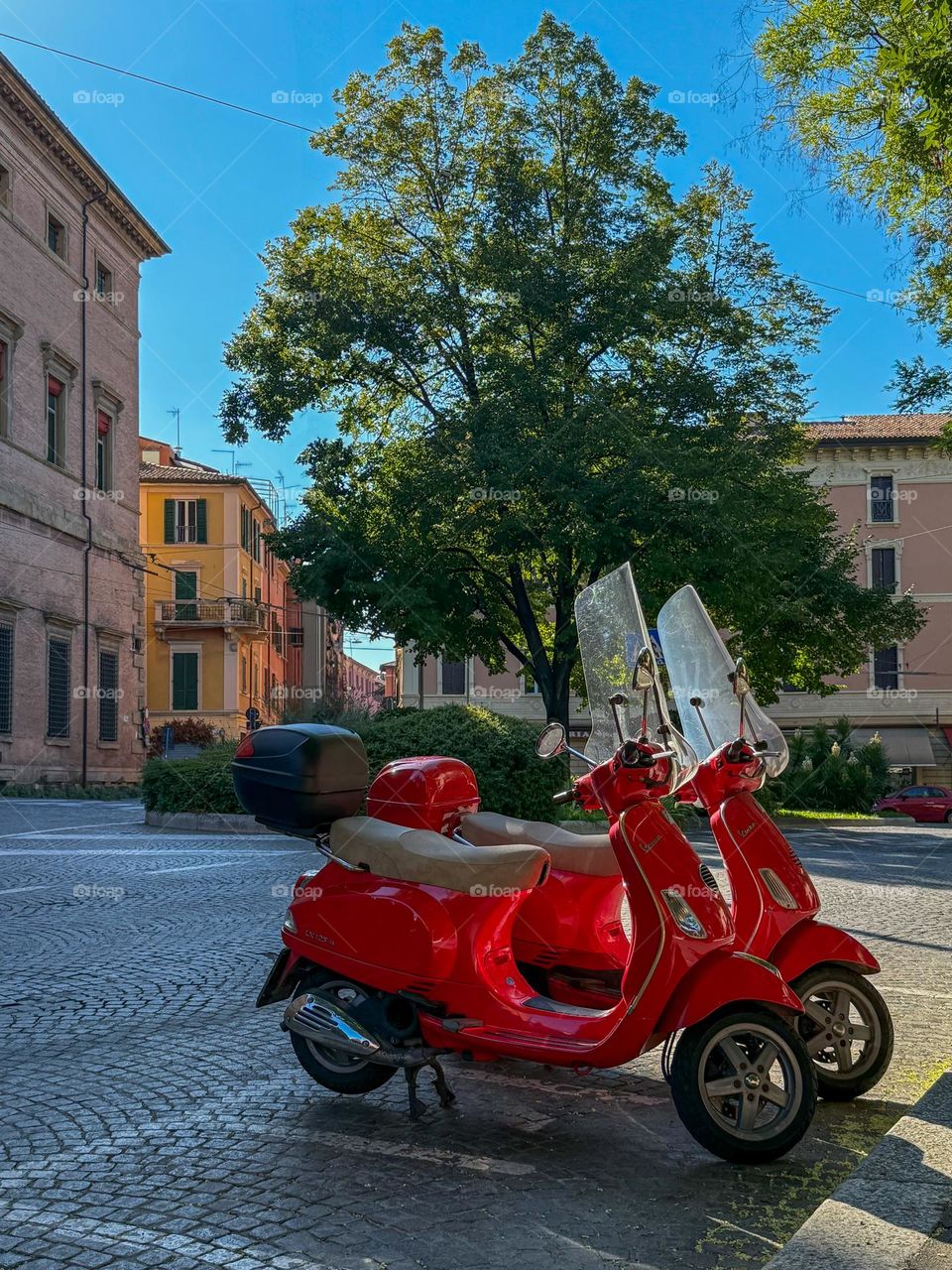 Two bright red scooters parked on a cobblestone street in a picturesque European town, Bologna, Italy, on a sunny summer  day, with colorful buildings and green trees in the background.