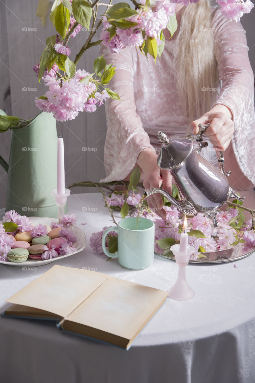young woman in a pink velvet dress pours coffee into a turquoise cup from a silver coffee pot against a background of pink sakura flowers