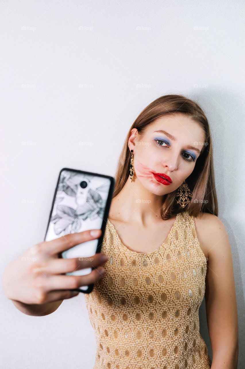 Pretty blonde girl with extreme make-up takes a photo of herself (selfie) with her phone; her lipstick is smeared but she looks confident