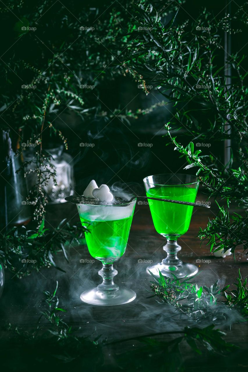 Two glasses of absinthe surrounded by wormwood stand on a dark wooden table and seem to glow from the inside. Tempting alcoholic beverages, mixology and bar art.