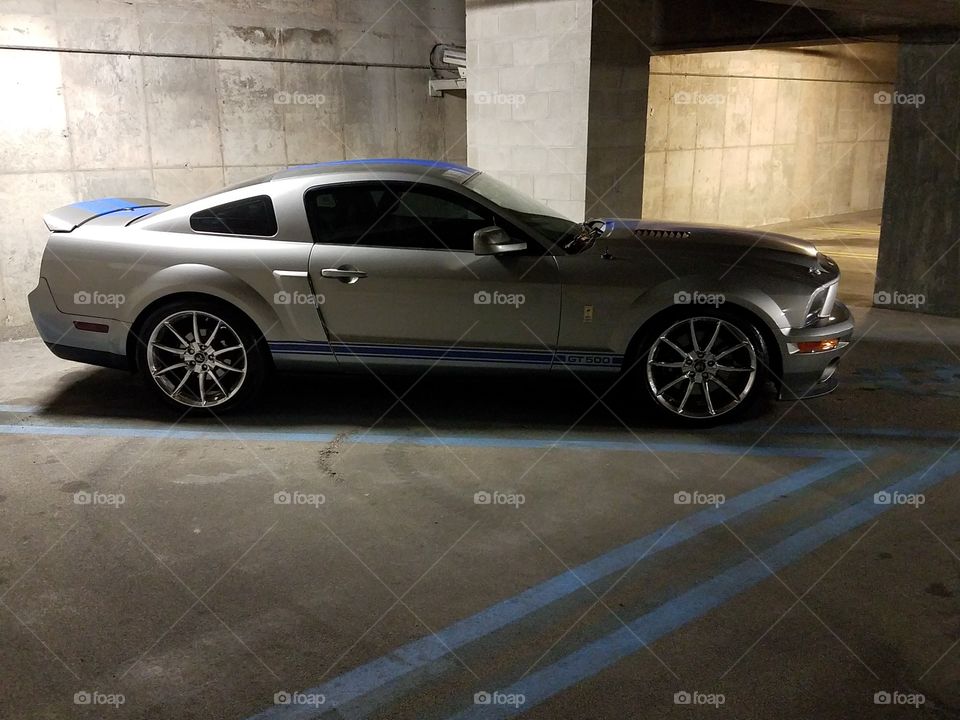 A silver Shelby mustang with blue racing stripes takes up a handicap spot in a garage.