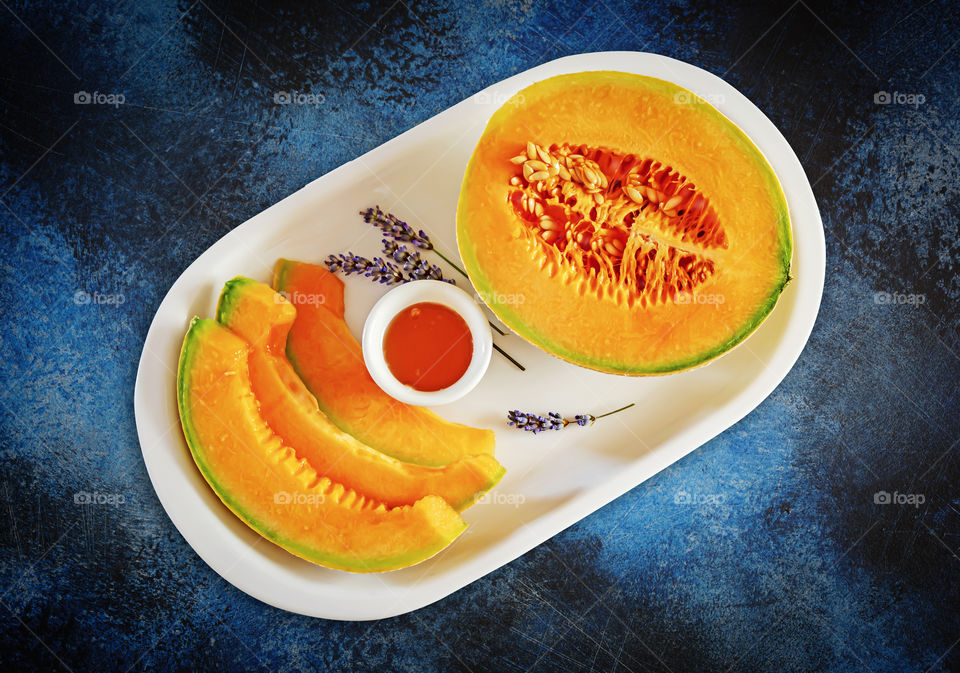 Concept of local, natural, seasonal fruits and vegetables. Sweet melon cut into slices with honey and lavender on a white plate on a blue textured background. Sweet healthy snack. Food photos