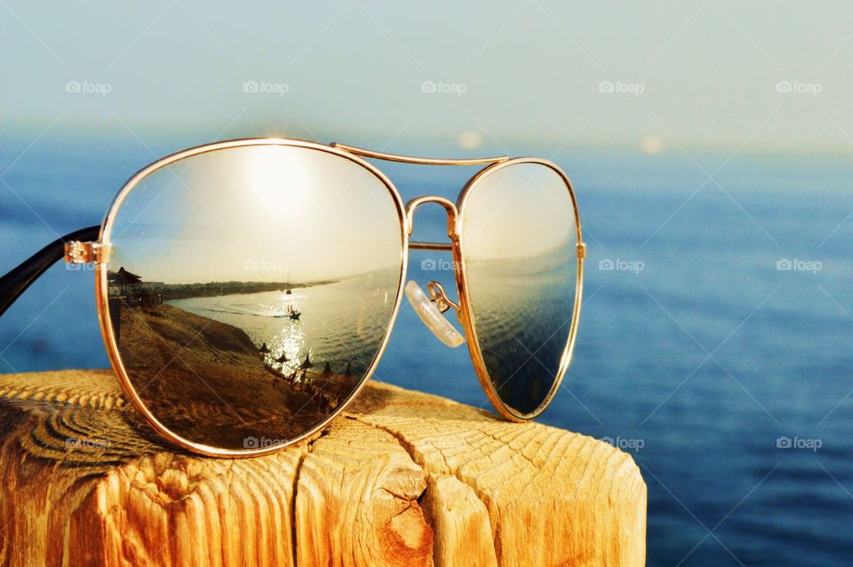 Eine Sonnenbrille auf einem Holzbalken, angestrahlt vom Sonnenuntergang. Diese reflektiert den Strand und ein Boot im Roten Meer.