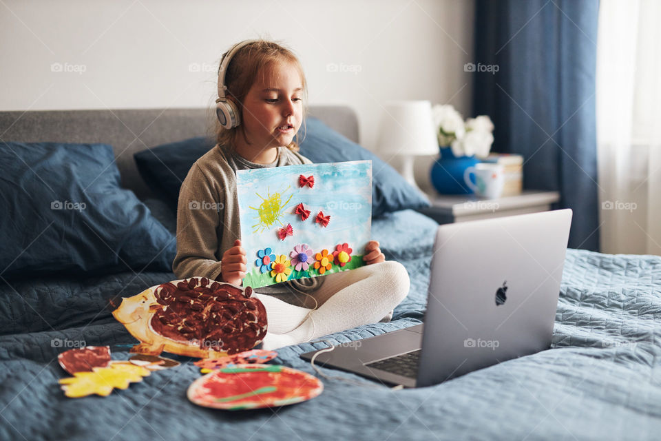 Using Apple MacBook.Little girl preschooler learning online showing her works drawings done at home. Child learning watching lesson remotely, talking with tutoress on video call from home during quarantine. Kid using laptop, headphones sitting on bed
