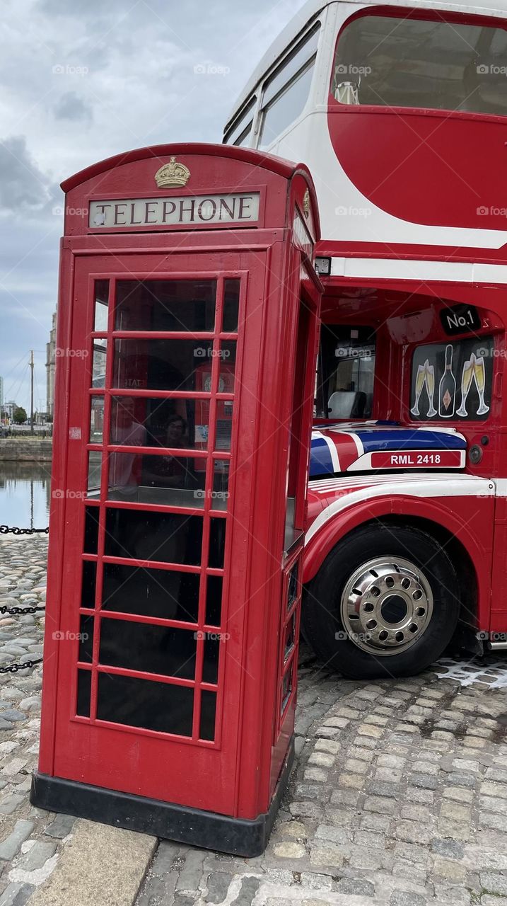 A classic scene from Liverpool, UK, featuring a bright red telephone box, a quintessential British phone kiosk popular in the 1940s and 1950s. Behind it is a red double-decker bus, evoking a sense of nostalgia for British heritage and city life.