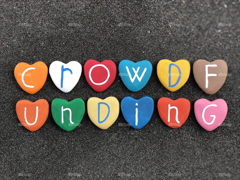 Crowdfunding text with multicolored heart stones over black volcanic sand