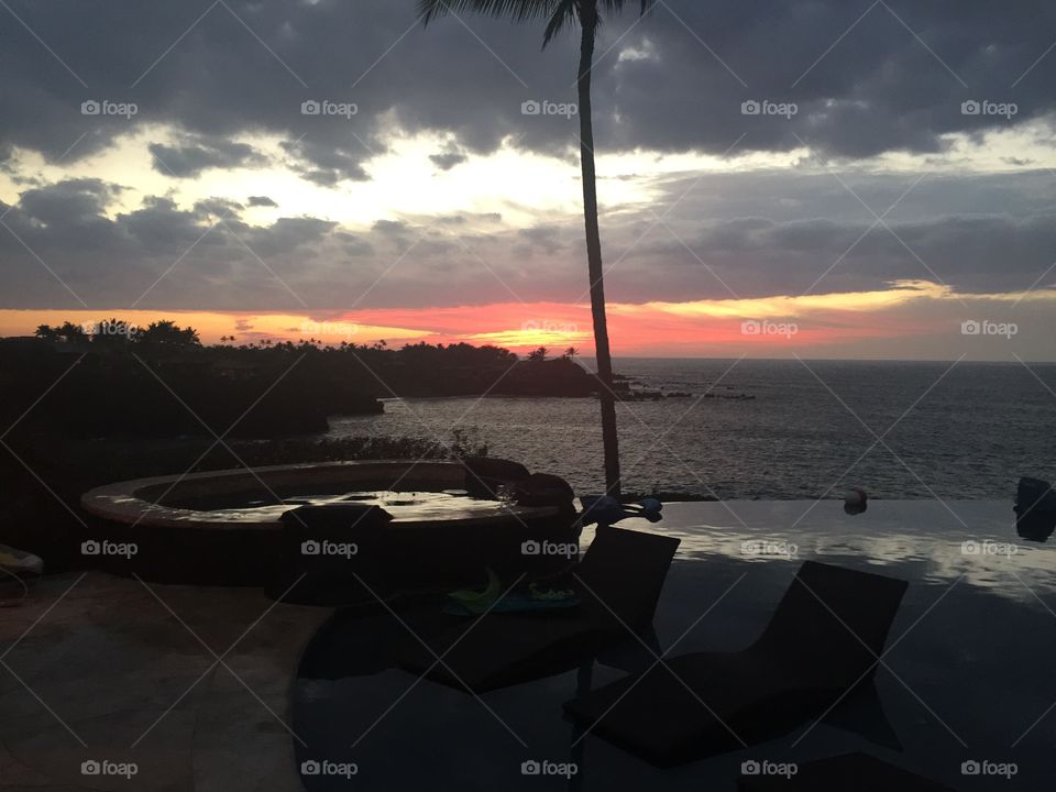 A cloudy sunset with a Rocky coastline.  A lone coconut palm tree in the center. Chaises in the foreground, on a pool deck where a pool and jacuzzi lie.