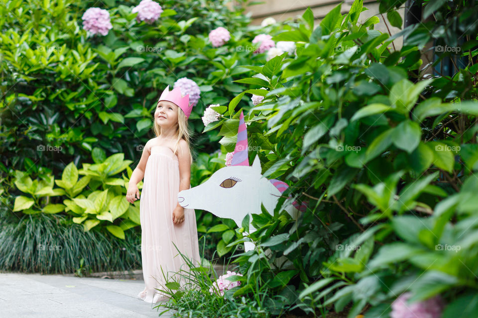 Happy little Caucasian girl with blonde hair and crown standing near unicorn in blooming garden at summer day