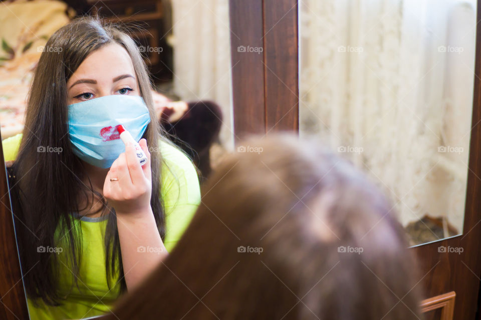 A young girl does makeup for a walk outside in a protective mask against the coronavirus pandemic, her lips are painted with red lipstick.