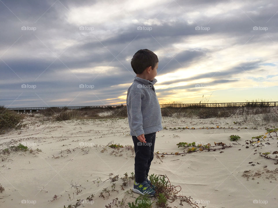 Child in sweater looking into distance on sandy beach at sunset with orange sky and pier