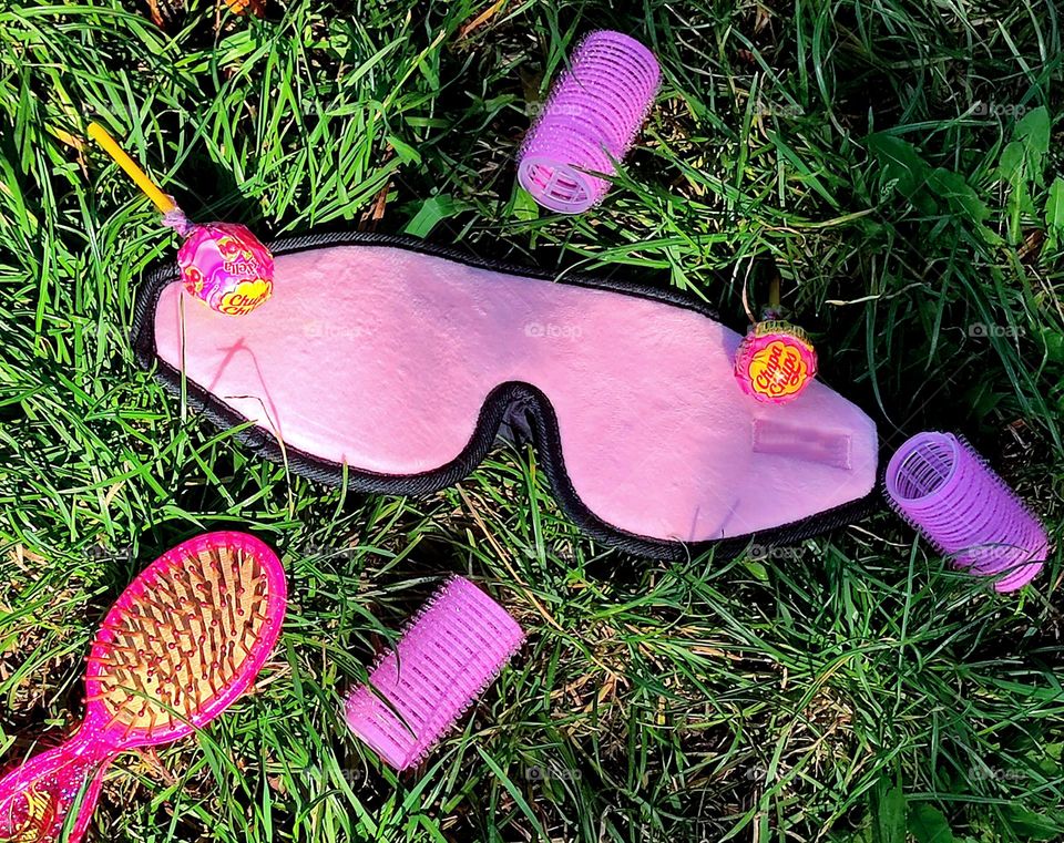 Barbie style.  On the green grass are: a pink eye mask, pink curlers, "Chupa Chyps" and a pink hairbrush