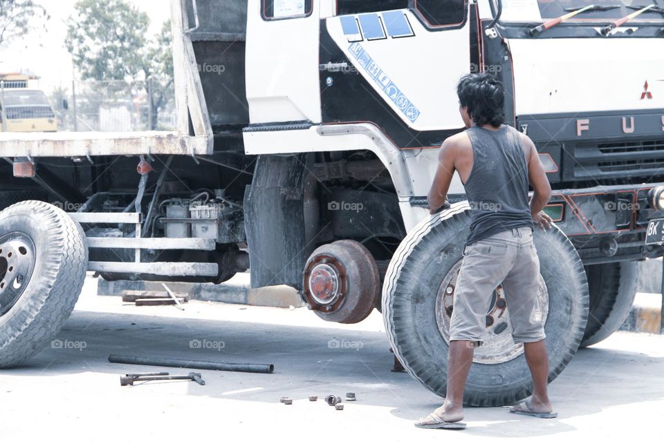 A large truck tire propped against the body of a truck by a man repairing it by the side of the road during the day.