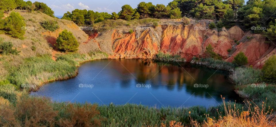 Beautiful natural background with a small lake formed in a quarry with red clay and greenery and trees growing around in Otranto, Italy, close-up side view. The concept of beautiful wallpaper, nature background, travel time.