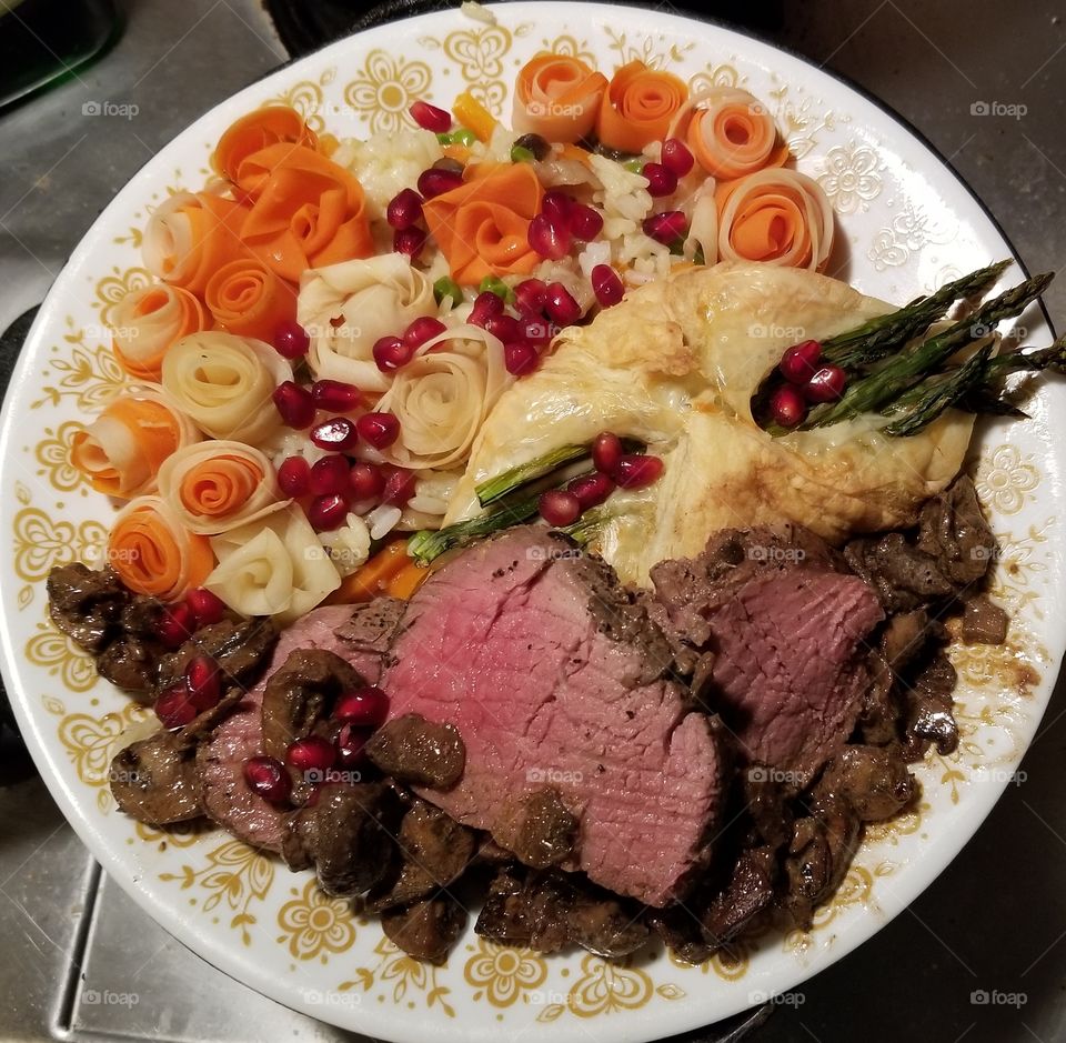Beef tenderloin, with mushrooms cooked in white wine, shallots, and sour cream; asparagus & gruyere wrapped in puff pastry; flowers made from ribbons of white and orange carrots, nestled in rice pilaf, and drizzled with butter, & Pomegranate arils.