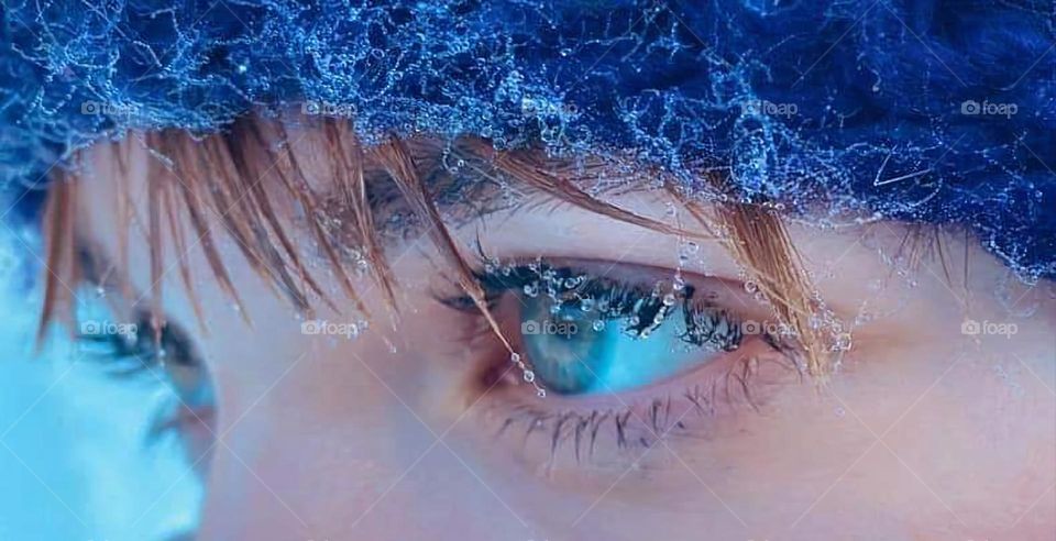 Close up on the blue eyes of a white ginger hair woman wearing a blue winter wool hat from which hair tips are sticking out. Her eye lashes, hair tips and wool hat are covered with frosty icy pearls.