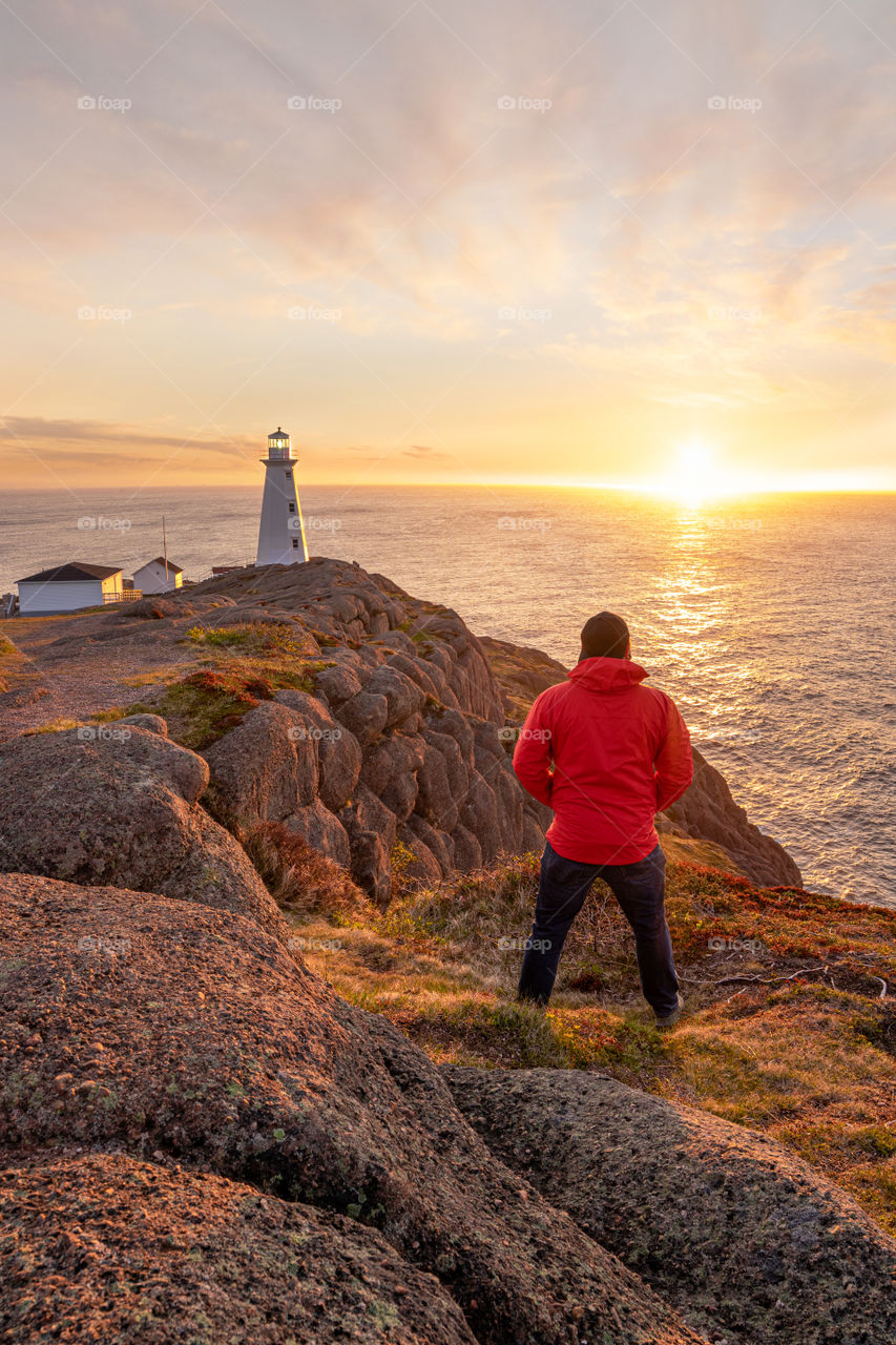 Young man in a red jacket standing on a rocky coastal cliff watching the sun rise over the ocean, with a white lighthouse on the edge of the shore. Cape Spear - Newfoundland 